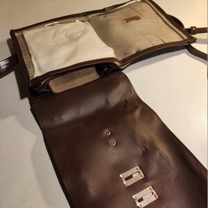 Puede incluir: Un bolso de mensajero de cuero marr&oacute;n con una solapa de pl&aacute;stico transparente. El bolso tiene una solapa con una ventana de pl&aacute;stico transparente y un cierre de cremallera. El bolso tiene una correa y herrajes met&aacute;licos.