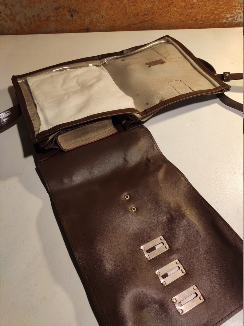 Puede incluir: Un bolso de mensajero de cuero marr&oacute;n con una solapa de pl&aacute;stico transparente. El bolso tiene una solapa con una ventana de pl&aacute;stico transparente y un cierre de cremallera. El bolso tiene una correa y herrajes met&aacute;licos.