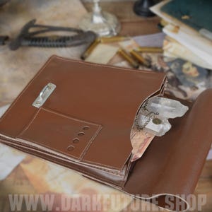 May include: A brown leather portfolio with a silver clasp and a small pocket. Inside, a clear quartz crystal hangs from a chain. The portfolio is open, revealing a map and other items.