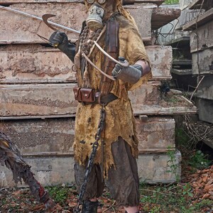 May include: A person wearing a brown, tattered, and worn costume with a gas mask and a spiked helmet. The person is holding a long, curved metal weapon. The person is standing in front of a pile of concrete blocks.