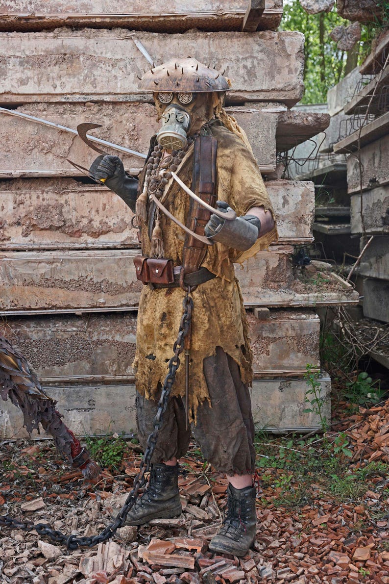 May include: A person wearing a brown, tattered, and worn costume with a gas mask and a spiked helmet. The person is holding a long, curved metal weapon. The person is standing in front of a pile of concrete blocks.