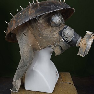 May include: A brown and black gas mask with a metal helmet with spikes. The helmet is covered in a brown fabric and has a clear plastic visor. The gas mask has a black rubber mouthpiece and a brown fabric filter.