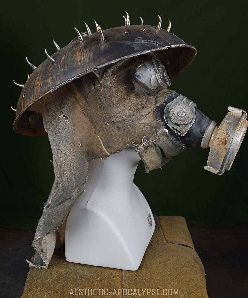 May include: A brown and black gas mask with a metal helmet with spikes. The helmet is covered in a brown fabric and has a clear plastic visor. The gas mask has a black rubber mouthpiece and a brown fabric filter.