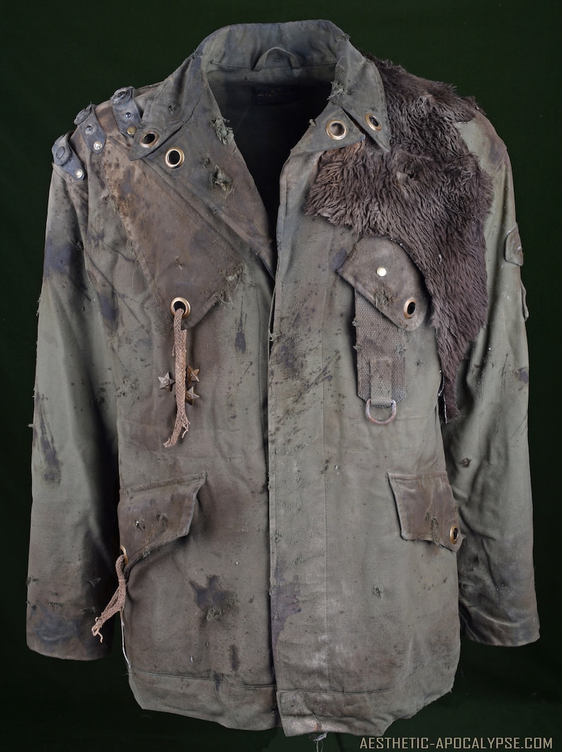 K&ouml;nnte beinhalten: Eine gr&uuml;ne, abgenutzte Canvas-Jacke mit einem Pelzkragen, Metallverzierungen und einem abgenutzten Lederriemen. Die Jacke hat einen abgenutzten Look mit Schmutz- und Abnutzungsspuren.