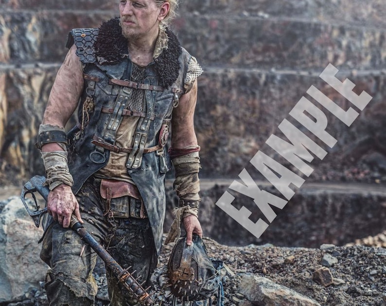 K&ouml;nnte beinhalten: Ein Mann tr&auml;gt eine dunkelbraune Lederweste mit mehreren Riemen und Schnallen, ein schwarzes Hemd und eine dunkelblaue Jeans. Er h&auml;lt einen Stachelschl&auml;ger in seiner linken Hand und einen Metallhelm in seiner rechten Hand. Der Hintergrund ist eine felsige, graue Landschaft.