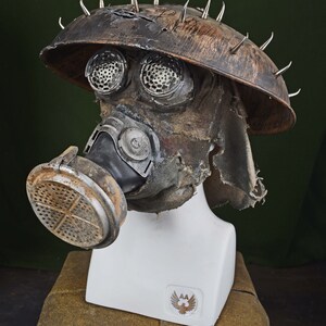 May include: A brown, weathered gas mask with a metal helmet and spikes. The mask has a black rubber mouthpiece and a filter on the side. The helmet has two round, metal eye pieces.