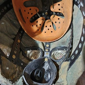 May include: A close-up of the inside of a gas mask. The mask is black and has a green fabric lining. The mask has two round filters on the sides and a round mouthpiece in the center. The mask is attached to a brown leather helmet with a strap and buckles.