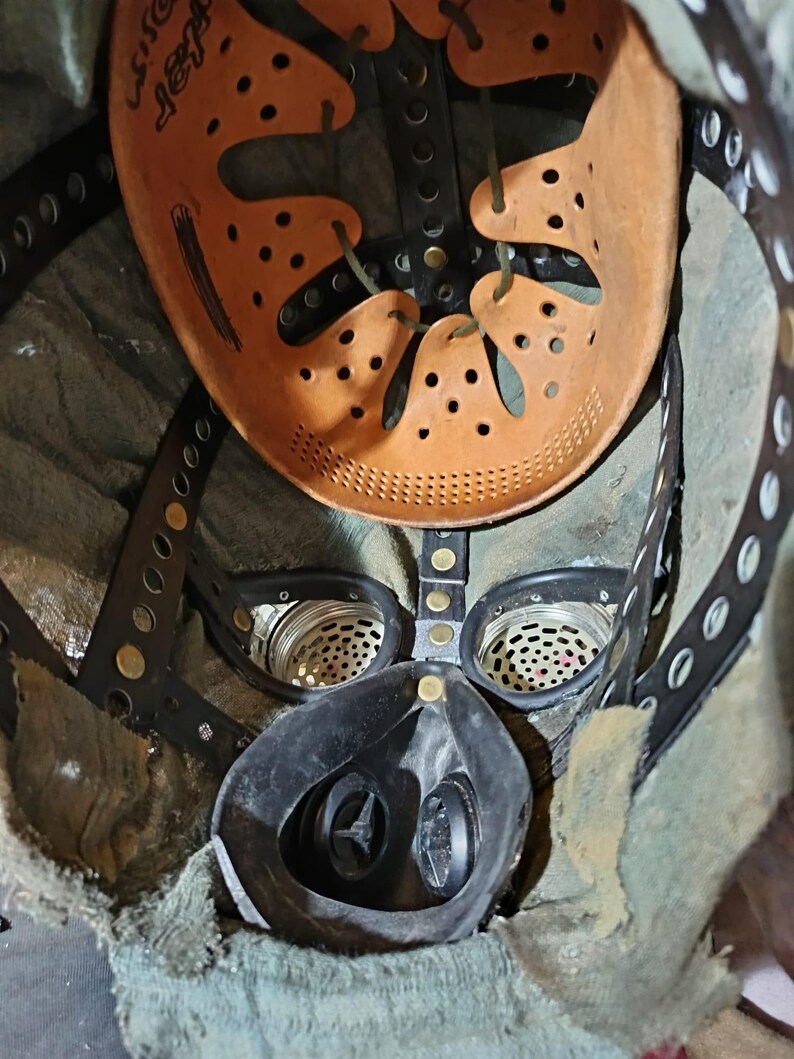 May include: A close-up of the inside of a gas mask. The mask is black and has a green fabric lining. The mask has two round filters on the sides and a round mouthpiece in the center. The mask is attached to a brown leather helmet with a strap and buckles.