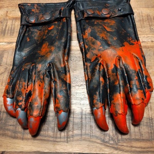 Op de afbeelding: Een paar zwarte leren handschoenen met oranje spatten verf en zilveren klauwen. De handschoenen hebben een zwarte band met twee zilveren knopen.