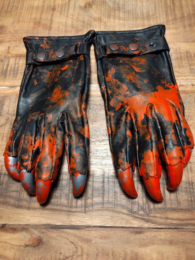 Op de afbeelding: Een paar zwarte leren handschoenen met oranje spatten verf en zilveren klauwen. De handschoenen hebben een zwarte band met twee zilveren knopen.