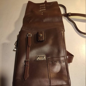 Puede incluir: Una cartera de cuero marr&oacute;n con cierre de solapa, una correa con hebilla y un peque&ntilde;o bolsillo en la parte delantera. La bolsa tiene una correa larga para llevarla al hombro.