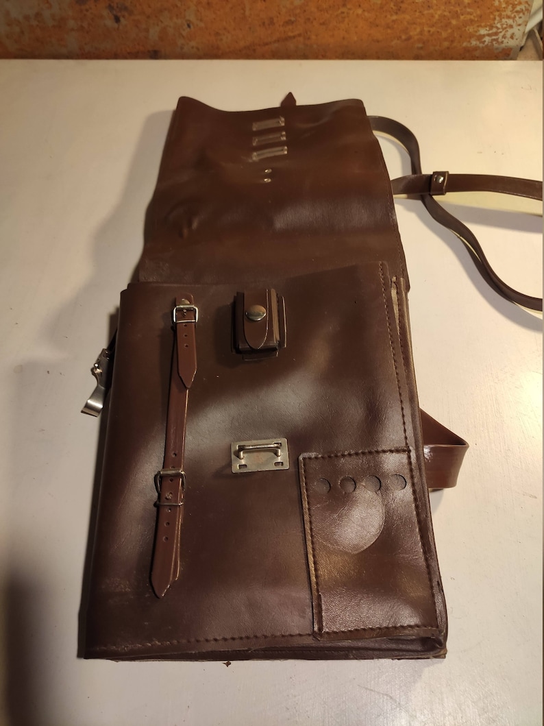 Puede incluir: Una cartera de cuero marr&oacute;n con cierre de solapa, una correa con hebilla y un peque&ntilde;o bolsillo en la parte delantera. La bolsa tiene una correa larga para llevarla al hombro.