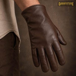 Può includere: Un guanto in pelle marrone scuro con una finitura liscia e un design semplice del polsino. Il guanto è mostrato in una posizione neutra con le dita estese. Nell'angolo in alto a destra, il testo DARKFUTURE è visibile in un riquadro giallo.