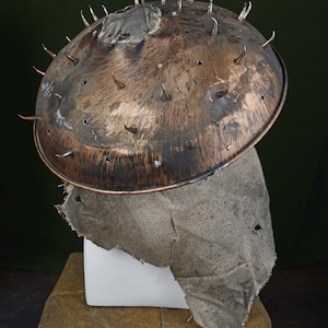 May include: A rusty metal helmet with spikes and a fabric covering. The helmet is on a white stand and a brown surface.