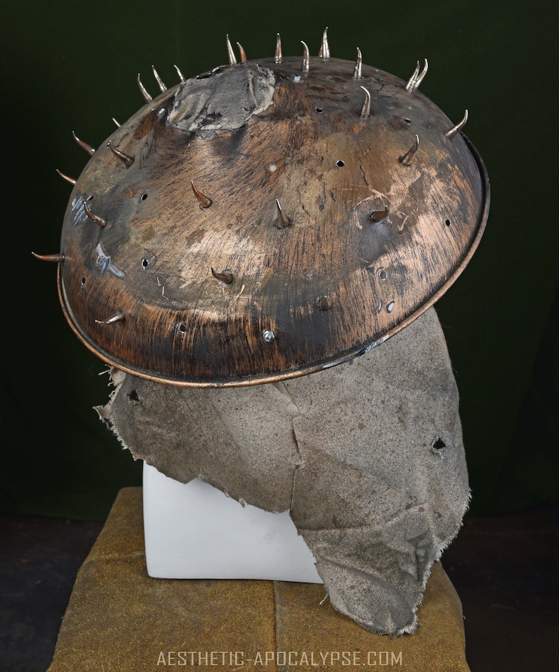 May include: A rusty metal helmet with spikes and a fabric covering. The helmet is on a white stand and a brown surface.