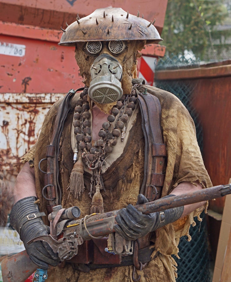 May include: A person wearing a tattered, brown, and beige costume with a gas mask and a large, rusty, metal rifle. The person is wearing a brown leather strap across their chest and a beaded necklace. The person is standing in front of a red metal container.
