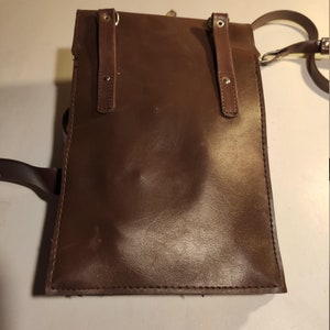 Puede incluir: Un bolso de cuero marr&oacute;n con dos correas ajustables y una correa larga para el hombro. El bolso tiene un cierre de solapa con dos hebillas.