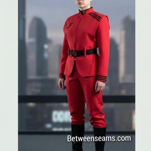 Può includere: Un completo uniforme rosso brillante composto da una giacca strutturata con colletto alto, finiture nere e una cintura nera con fibbia circolare. La giacca presenta strisce nere sulle spalle e sui polsini, e un logo nero sul petto e sul colletto.
