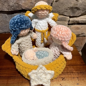 Puede incluir: Un belén de ganchillo con un pesebre amarillo, un ángel blanco, un ángel azul, un ángel rosa y un bebé Jesús. El pesebre está decorado con una estrella blanca.