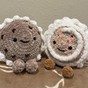 May include: Two plush, round, crocheted toys resembling cookies. One is brown with chocolate chip details, the other is pink with colorful sprinkles. Both have white frosting edges and smiling faces. Small, round, brown crocheted balls are also present.