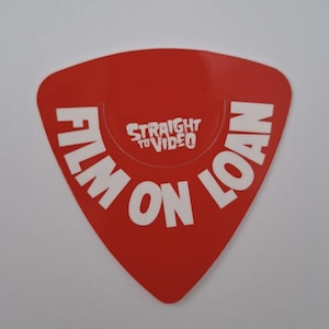 May include: A red, guitar pick-shaped sticker with the words "FILM ON LOAN" in white, bold, capital letters. The text "STRAIGHT TO VIDEO" is in a smaller font in the centre.