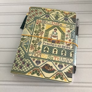May include: A green and cream fabric-covered journal with a yellow rubber band and a green tassel. The cover features a cross-stitch design with the text "Peaceful is my garden".