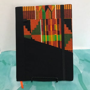 May include: A black notebook cover with a zippered pocket and a black elastic band closure. The cover is decorated with a colorful African print fabric featuring geometric patterns in shades of orange, green, and brown.