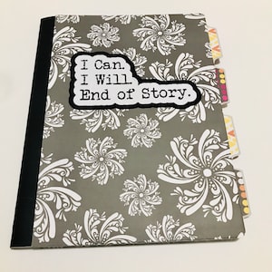 May include: A gray notebook with a white floral pattern and black spine. The front cover has a white speech bubble with the text "I Can. I Will. End of Story."