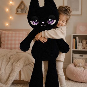 Könnte beinhalten: Ein großes, schwarzes, gehäkeltes Plüschtier in Form einer Katze mit langen Ohren, einer kleinen rosa Nase und zwei kleinen weißen Fangzähnen. Die Katze hat große, ausdrucksstarke violette Augen mit weißen Glanzlichtern und eine weiche Textur.