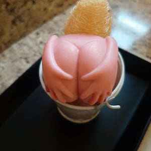 Puede incluir: Un jabón rosa con forma de manos que sostiene una pequeña esponja amarilla esponjosa en un cubo blanco.