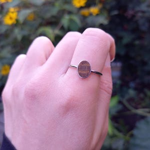 Puede incluir: Un anillo de plata con una piedra central ovalada con un patrón de vetas de madera. El anillo se encuentra sobre un fondo de follaje verde y flores amarillas. Una joya sencilla y elegante.