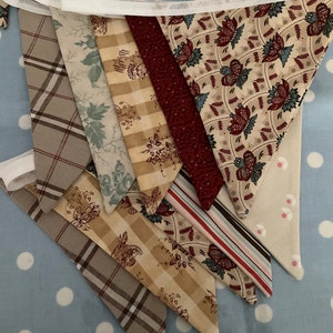 May include: A decorative bunting featuring triangular fabric flags in a variety of patterns. The fabrics include plaid, floral, and striped designs in beige, brown, red, and blue. The pennants are attached to a white ribbon.