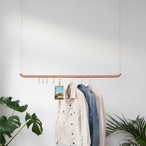 May include: A copper-colored metal clothing rack suspended from the ceiling by two light-colored ropes. The horizontal bar features several metal hooks, one holding a small photograph, and three metal hangers with jackets hanging from them.