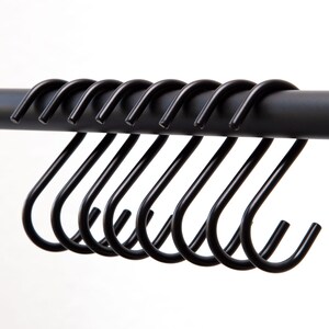 Puede incluir: Primer plano de una fila de ganchos en S de metal negro colgados de una barra de metal negro. Los ganchos están espaciados uniformemente y tienen un diseño suave y curvado. El fondo es blanco liso.