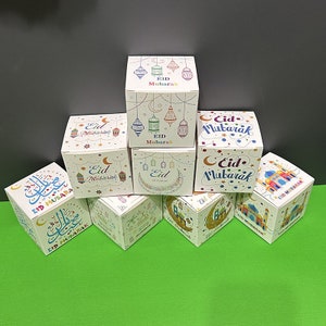 May include: Assortment of small white gift boxes with colorful designs and the text "Eid Mubarak" in Arabic and English. The boxes feature various Islamic motifs, including crescent moons, stars, and lanterns.