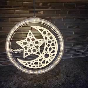 May include: A white illuminated crescent moon and star design with an intricate geometric pattern. The design is surrounded by a clear plastic ring with a string hanging from the top. The words "Ramadan Kareem" are written in white on the design.