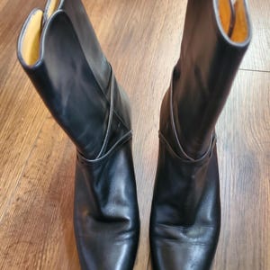 May include: A pair of black leather boots with a mid-height heel. The boots have a smooth, polished finish and a folded-over top revealing a yellow interior. The boots are displayed on a wooden surface.
