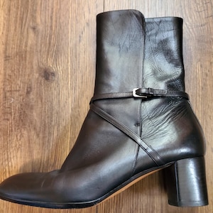 May include: Black leather ankle boot with a block heel and a strap detail around the ankle. The boot has a rounded toe and a stacked heel. The heel height is approximately 3 inches.