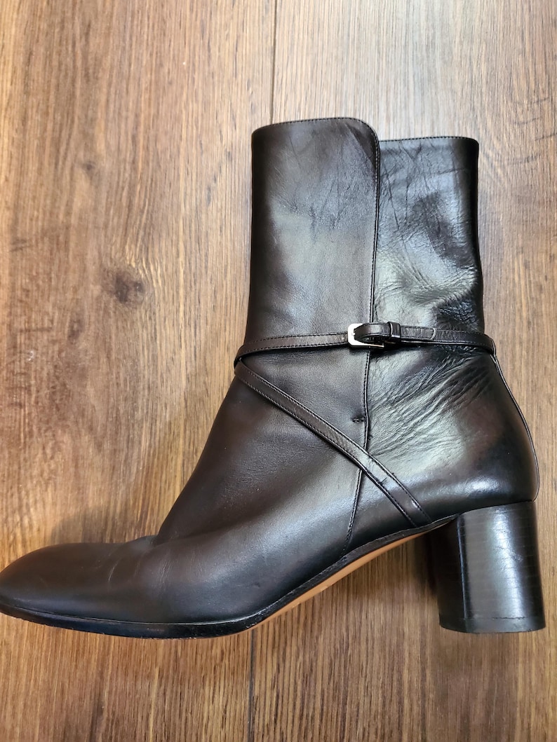 May include: Black leather ankle boot with a block heel and a strap detail around the ankle. The boot has a rounded toe and a stacked heel. The heel height is approximately 3 inches.