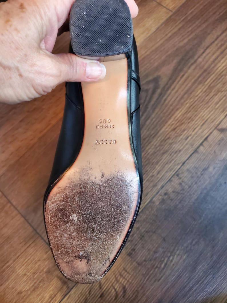 May include: Black leather shoe with a worn sole. The shoe has a block heel and the brand name "BALLY" is visible. The sole shows signs of wear and tear, indicating previous use. The shoe size is US 6 and EU 36 1/2.
