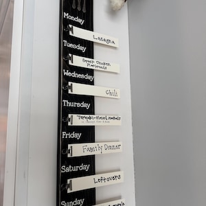 Puede incluir: Un tablero de menú de madera negra con texto blanco y clips metálicos. El tablero tiene los días de la semana enumerados con una comida diferente para cada día. Las comidas incluyen Lasagna, Flatbreads de pollo griego, Chili, Albóndigas glaseadas con teriyaki, Cena familiar, Sobras y Cazuela de fideos de atún.