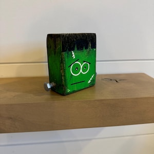 Könnte beinhalten: Ein grüner Holzblock mit einem bemalten Gesicht von Frankensteins Monster. Der Block hat schwarze Haare, grüne Haut, weiße Nähte und grüne Augen. Der Block ist mit zwei silbernen Schrauben an einer Holzoberfläche befestigt.