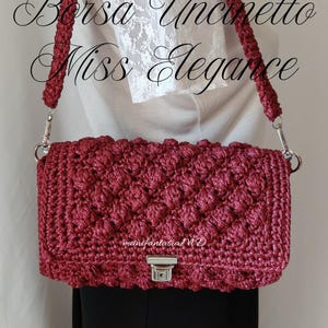 May include: A burgundy crochet handbag with a textured pattern and silver-tone hardware. The bag has a matching braided strap and a rectangular shape. Text on the bag reads "Borsa Uncinetto Miss Elegance" and "manifantasia MD".