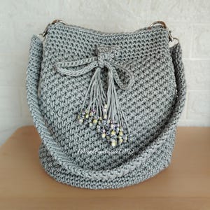 Può includere: Una borsa a secchiello in crochet grigia con chiusura a coulisse e un pendente a forma di nappa con perline. La borsa ha una tracolla lunga per essere portata a tracolla.