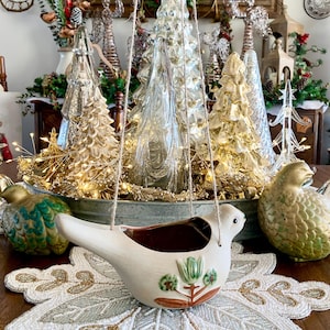 May include: A white ceramic bird-shaped hanging planter with a brown interior and floral design. The planter hangs from twine, with gold and silver Christmas tree decorations in the background. Two decorative bird figurines are on either side.