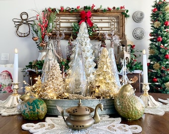 May include: A festive Christmas centerpiece featuring glass Christmas trees in gold and silver tones, fairy lights, and decorative birds. The arrangement includes a vintage brass teapot and lit candles, creating a warm holiday ambiance.