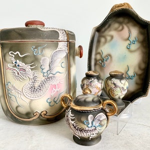 May include: A set of four vintage ceramic dragon-themed serving pieces. The set includes a large, lidded container, a small serving dish, and two small salt and pepper shakers. The pieces are decorated with a white dragon design on a cream background with gold accents.