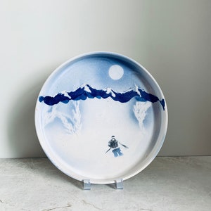 Puede incluir: Un plato de cerámica blanco con una escena de invierno azul y blanco. La escena representa a un esquiador en primer plano, montañas en el fondo y una luna llena en el cielo.