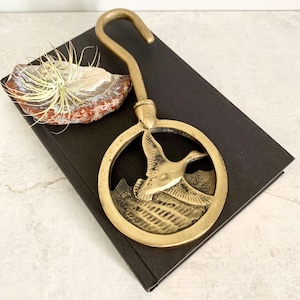 May include: A vintage brass duck-themed bottle opener with a hook. The opener features a circular design with a flying duck silhouette. The opener is on a black surface, next to a decorative rock with a plant.