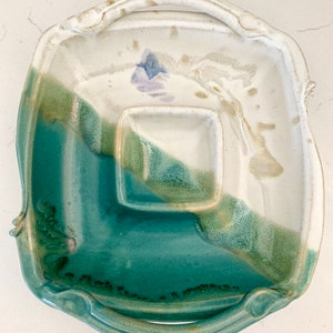 May include: Handmade ceramic serving platter with a square shape and a built-in square dip bowl. The platter features a teal, green, and white glaze with a floral accent. It has handles on each side.